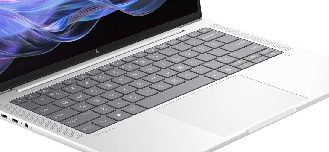 Zbliżenie na klawiaturę laptopa HP EliteBook w kolorze srebrnym, widoczne szare klawisze z białymi oznaczeniami, po bokach perforowane kratki głośników, poniżej duży touchpad, fragment ekranu z kolorową grafiką w górnej części. - klawiatura HP Premium z białym podświetleniem