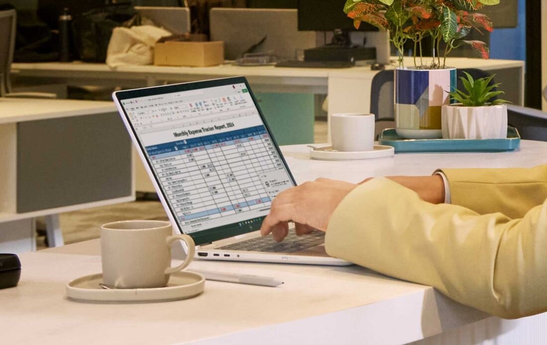 Osoba w żółtej marynarce pracuje na srebrnym laptopie HP przy okrągłym stole w biurze, na ekranie otwarty arkusz kalkulacyjny, obok filiżanka na spodku, długopis i doniczka z rośliną; w tle biurowe wnętrze. - Pamięć RAM LPDDR5X, dysk SSD PCIe NVMe 