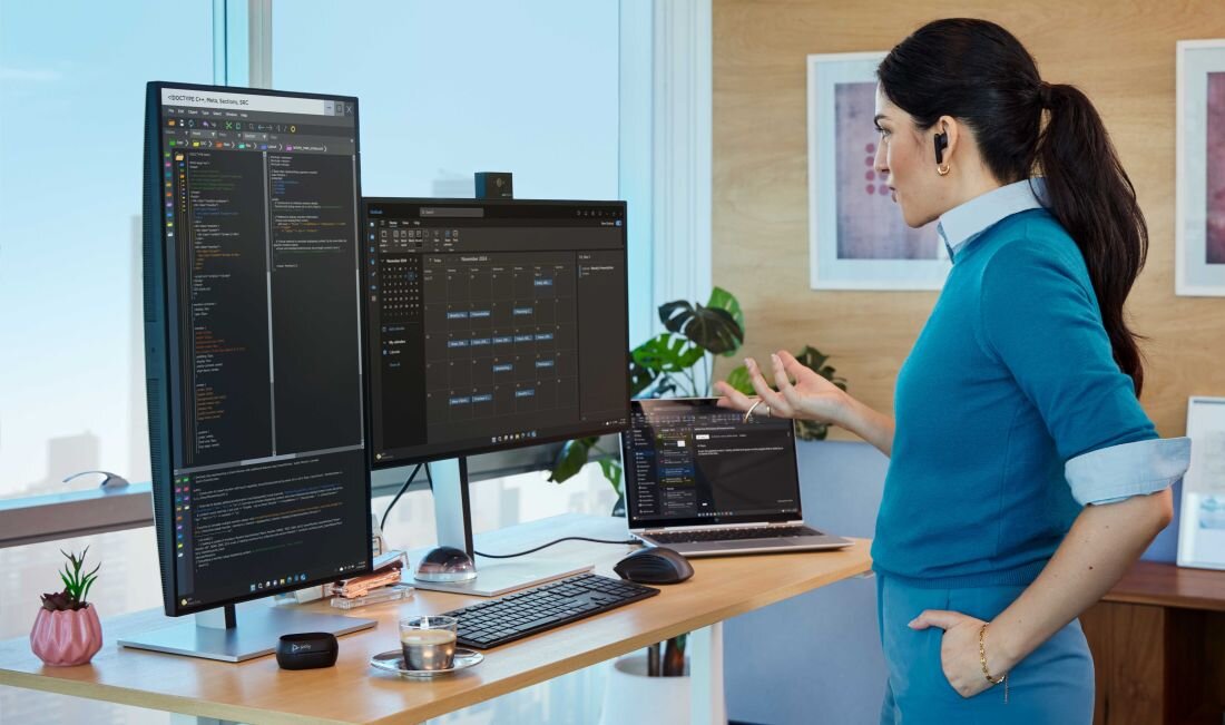 Kobieta w niebieskim swetrze stoi przy biurku z dużym monitorem, drugim monitorem poziomym oraz otwartym laptopem HP; na ekranach widoczne są aplikacje programistyczne i narzędzia do zarządzania projektami, na biurku klawiatura, myszka, filiżanka oraz rośliny doniczkowe, w tle nowoczesne, jasne biuro z dużymi oknami. - HP Sure Click, HP Sure Start