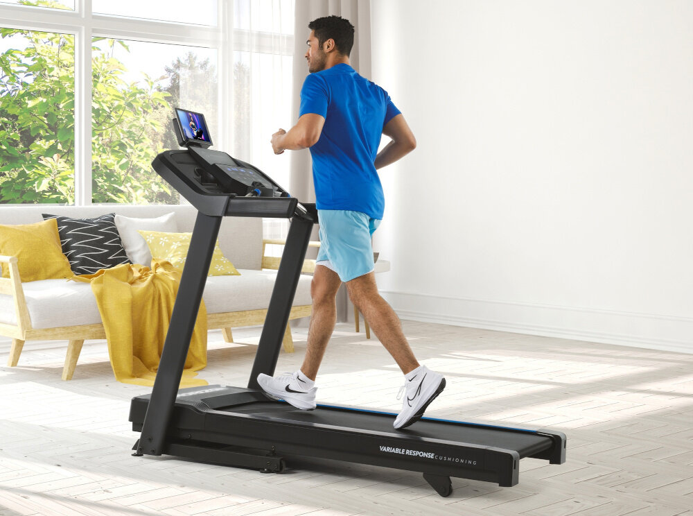 Bieżnia elektryczna HORIZON 5.0AT-21 1864W 16km-h pokój duże okno w tle kanapa pośrodku bieżnia na bieżni biegnie mężczyzna widok z boku trening w domowym zaciszu codzienna aktywność fizyczna zdrowie bieganie
