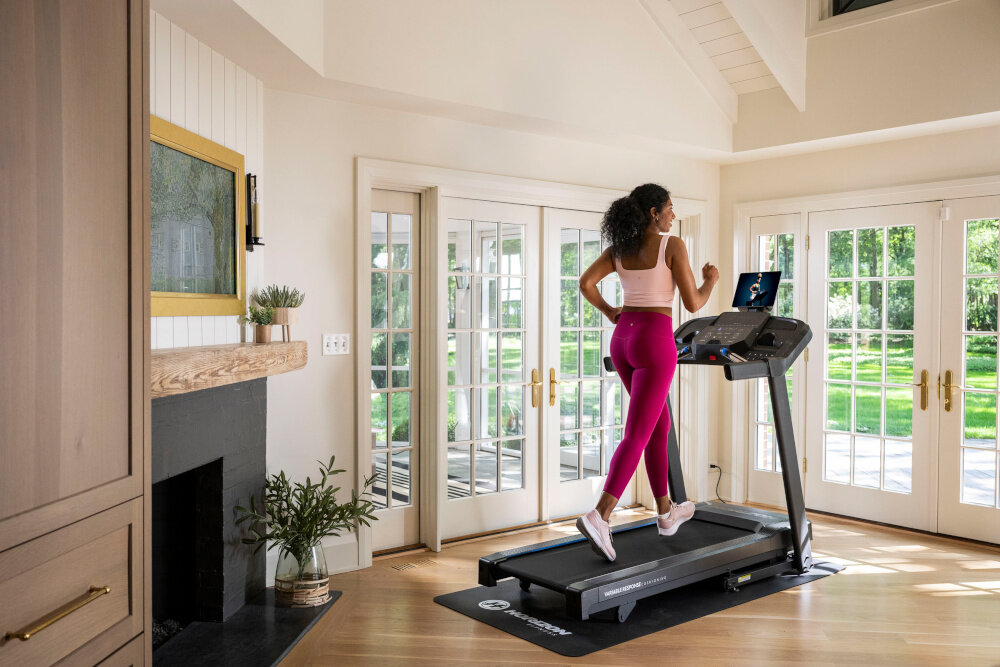 Bieżnia elektryczna HORIZON 5.0AT-21 1864W 16km-h pokój duże okna pośrodku bieżnia na bieżni biegnie kobieta widok od tyłu do użytku domowego utrzymanie dobrej kondycji kontrola masy ciała do marszu truchtu lub biegania dla użytkowników na różnych poziomach zaawansowania