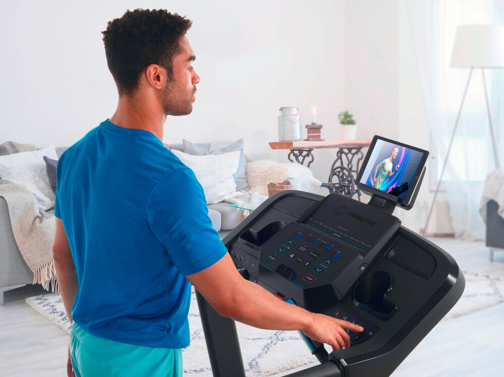 Bieżnia elektryczna HORIZON 5.0AT-21 1864W 16km-h pokój w tle kanapa mężczyzna na bieżni na uchwycie tablez z włączoną aplikacją łączność przez Bluetooth 4.0 integracja bieżni z aplikacją Horizon @Zone korzystanie z dodatkowych funkcji i zapis danych treningowych
