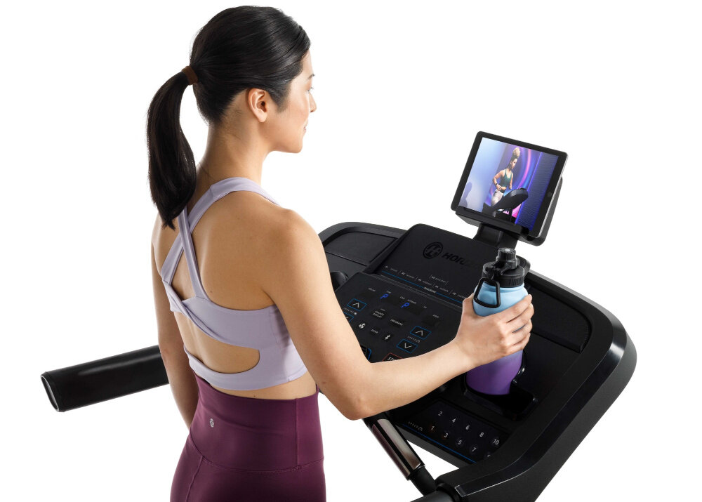 Bieżnia elektryczna HORIZON 5.0AT-21 1864W 16km-h kobieta stoi na bieżni widok na wyświetlacz kobieta stawia bidon w wyznaczonym miejscu zdjęcie na białym tle wchwyt na bidon miejsce na tablet komfort treningu możliwość oglądania filmów lub korzystania z aplikacji hydratacja jest na wyciągnięcie ręki ergonomiczne rozwiązania różnorodne funkcje