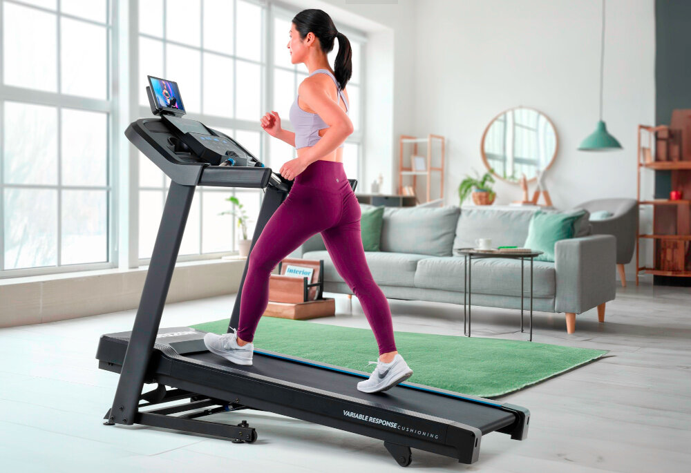 Bieżnia elektryczna HORIZON 5.0AT-21 1864W 16km-h kobieta biegnie na bieżni podniesiony pas biegowy pokój w tle kanapa bieżnia klasy H trwałość funkcjonalność waga 81,5 kg stabilność wysoka jakość materiałów przemyślana konstrukcja bezpieczeństwo solidne narzędzie do codziennego ruchu