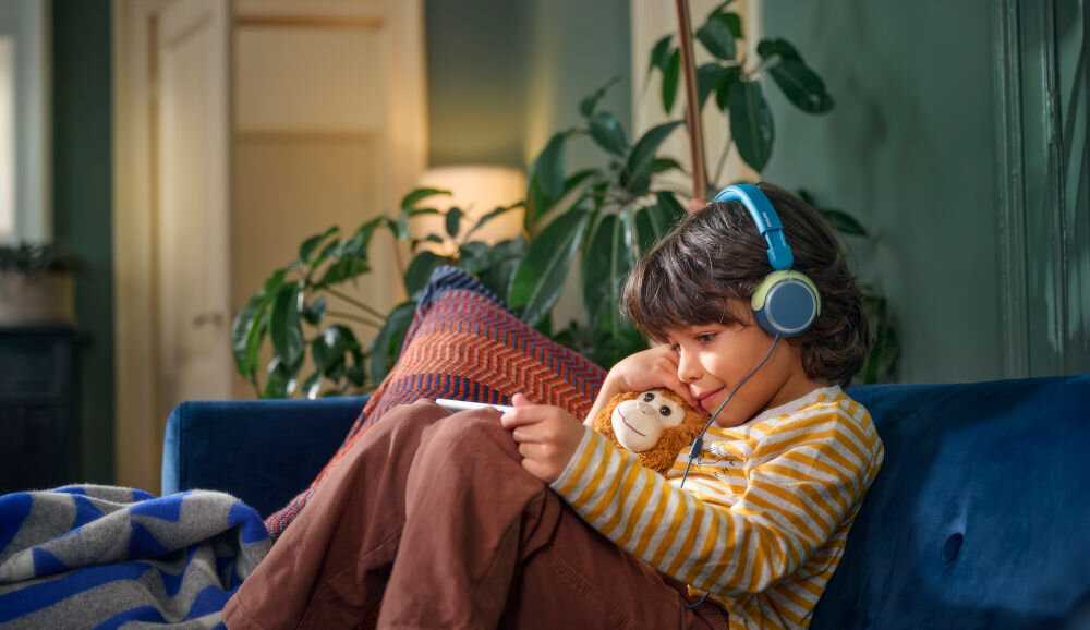 Słuchawki nauszne PHILIPS TAK2000MP miękkie poduszki i wygodny pałąk dla dzieci, dziecko w słuchawkach nausznych siedzące na kanapie z tabletem w ręku