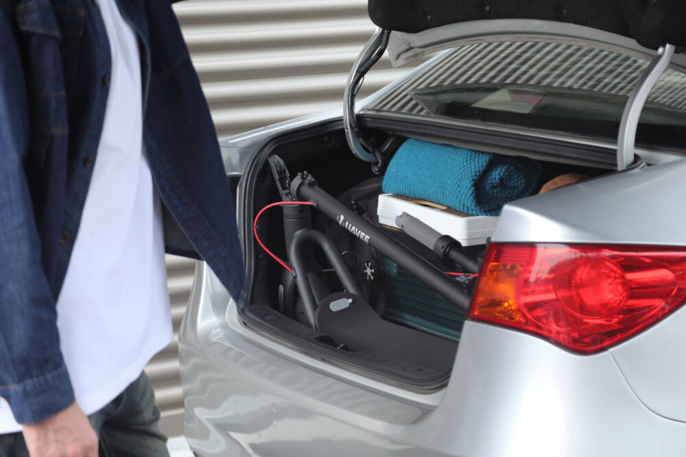 Hulajnoga elektryczna NAVEE V50i Pro 50km 350W 10 cali Czarny osoba stoi przy samochodzie otwarty bagażnik w bagażniku złożona hulajnoga składana konstrukcja innowacyjny system składania DoubleFlit zaoszczędzenie ponad 66% miejsca złożenie kierownicy do podłoża obrót kierownicy ułożenie wzdłuż konstrukcji