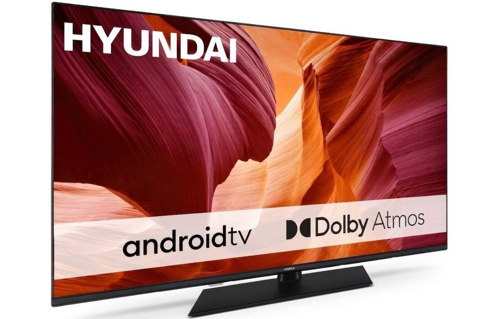 Telewizor z logo HYUNDAI na ekranie, wyświetlający abstrakcyjny obraz w odcieniach czerwieni i pomarańczu. Na ekranie widoczne są napisy: \'androidtv\' i \'Dolby Atmos\'.
