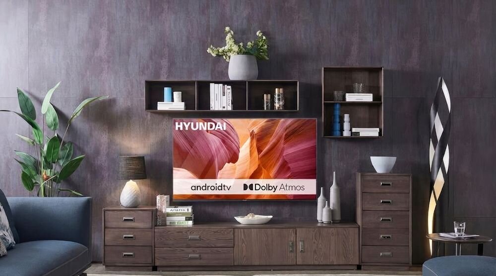W salonie na ścianie wisi telewizor z logo HYUNDAI, wyświetlający kolorową grafikę. Na ekranie widoczne są napisy: \'androidtv\', \'Dolby Atmos\'.