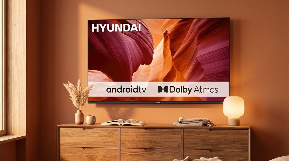Telewizor z logo HYUNDAI wyświetla obraz z napisami \'android tv\' i \'Dolby Atmos\'. W tle znajduje się drewniana komoda z dekoracjami i lampką.