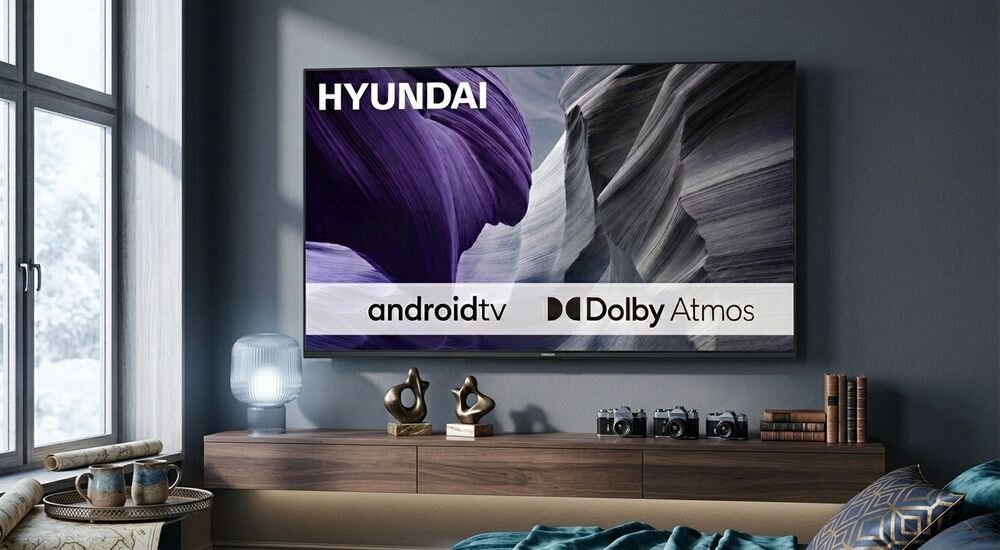 Telewizor na ścianie z wyświetlonym logo HYUNDAI oraz napisami androidtv i Dolby Atmos. W tle widoczny jest nowoczesny salon z drewnianą szafką i dekoracjami.