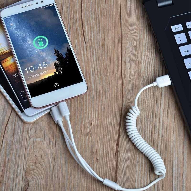 Na drewnianym blacie leży smartfon z wyświetlaczem pokazującym godzinę 10:45 i ikonę ładowania baterii. Telefon jest podłączony do laptopa za pomocą białego kabla USB z trzema różnymi końcówkami.
