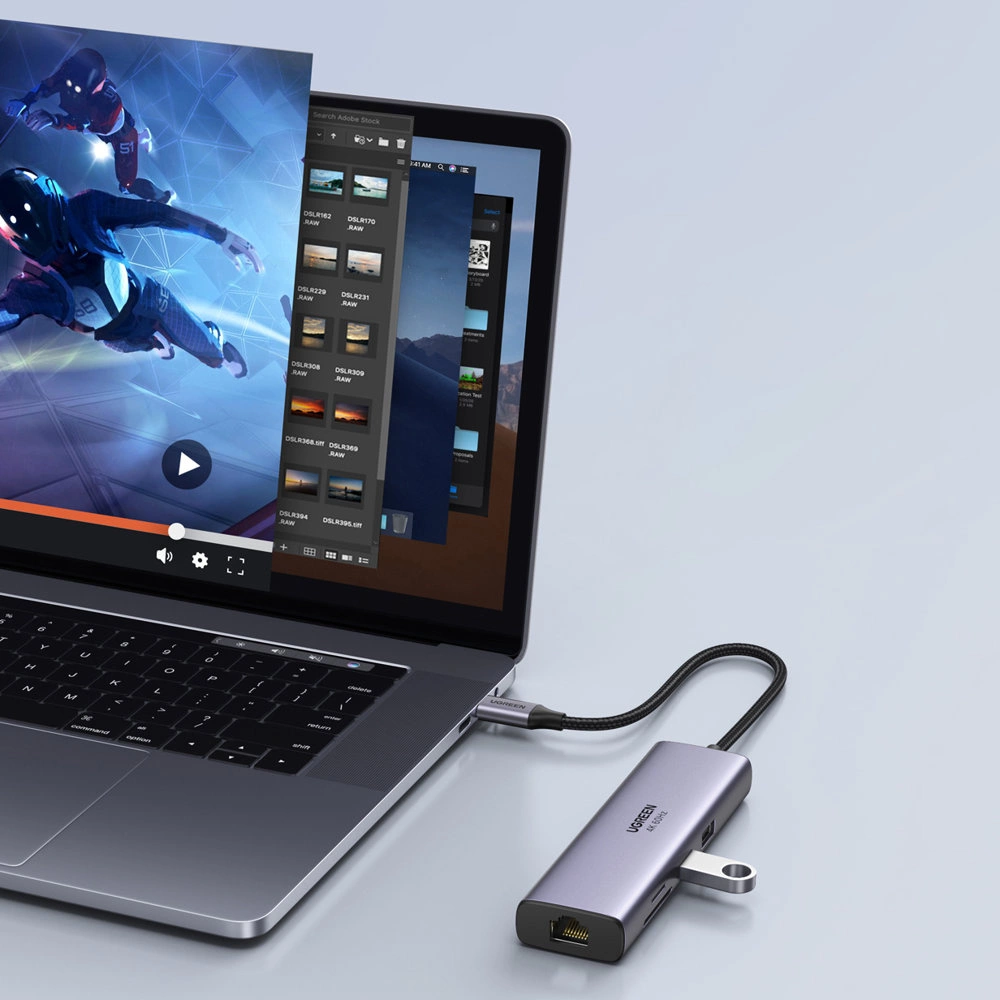 Na obrazie widać laptop z otwartym ekranem, na którym wyświetlane są różne okna aplikacji. Do laptopa podłączony jest szary hub Ugreen z kilkoma portami, w tym HDMI i USB-C, oraz widocznym logo \'UGREEN\'.