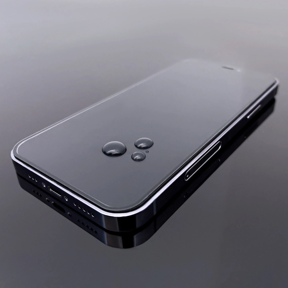 Na obrazie widoczny jest smartfon z przezroczystą folią ochronną na ekranie, na której znajdują się krople wody. Telefon leży na ciemnej, odbijającej powierzchni.