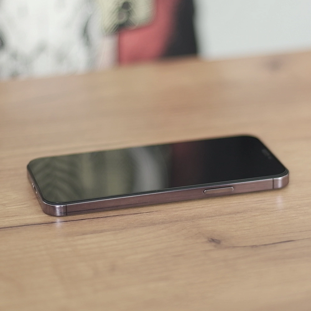 Na drewnianym stole leży smartfon z ciemnym ekranem skierowanym do góry. Obudowa telefonu jest metaliczna, a na boku widoczny jest przycisk.