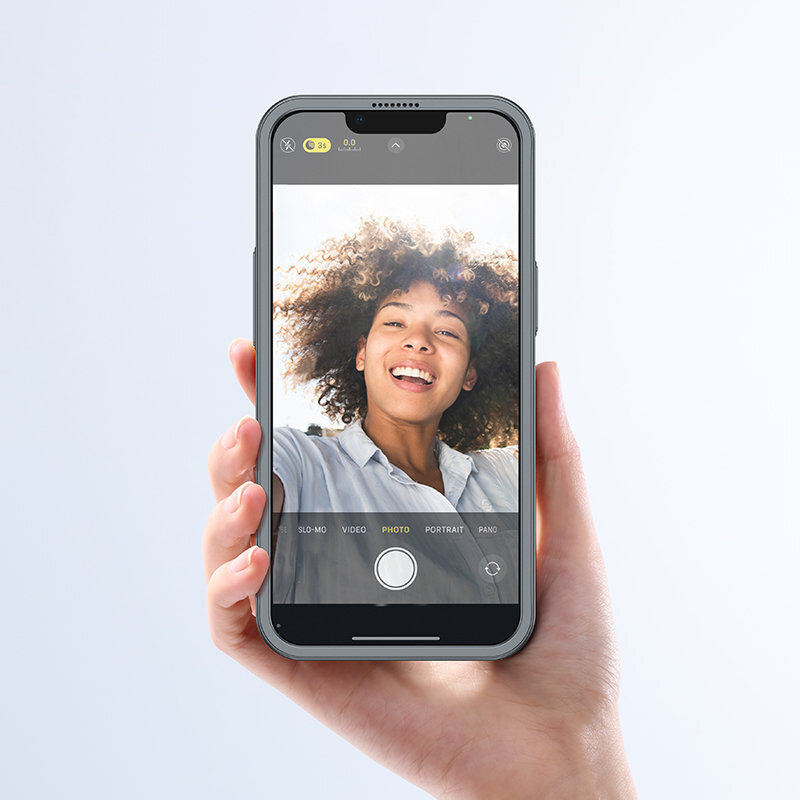 Osoba trzyma smartfon z włączoną aplikacją aparatu, na ekranie widoczna jest selfie uśmiechniętej osoby. Telefon jest w szarym etui, a na ekranie widoczne są opcje: SLO-MO, VIDEO, PHOTO, PORTRAIT, PANO.