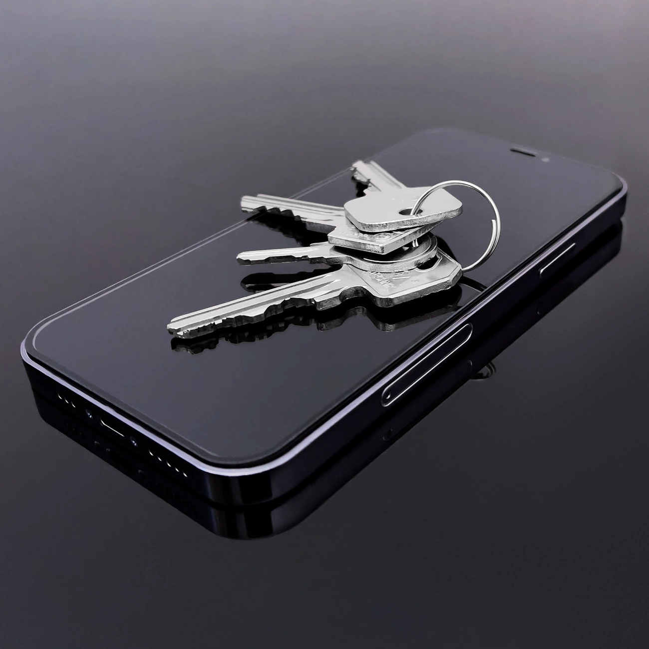 Na obrazie widać smartfon z czarną ramką, na którego ekranie leży kilka metalowych kluczy. Powierzchnia ekranu jest gładka i odbija światło.