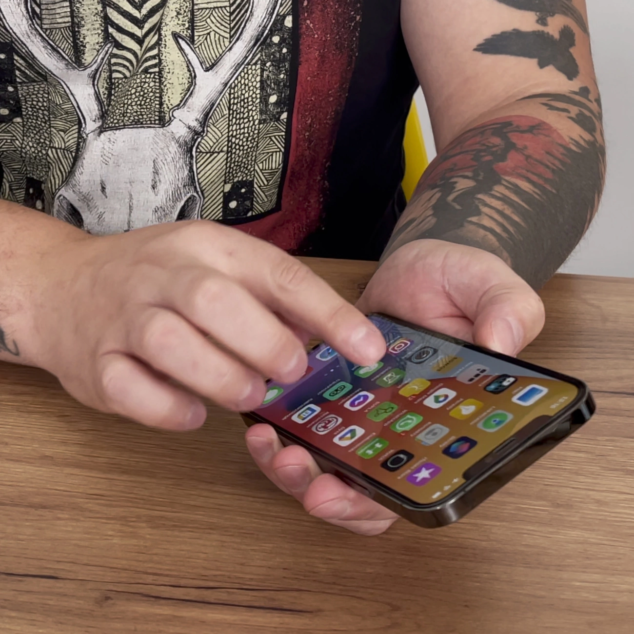 Osoba trzyma smartfon z widocznym ekranem pełnym kolorowych ikon aplikacji. Na rękach widoczne są tatuaże, a w tle znajduje się drewniany stół.