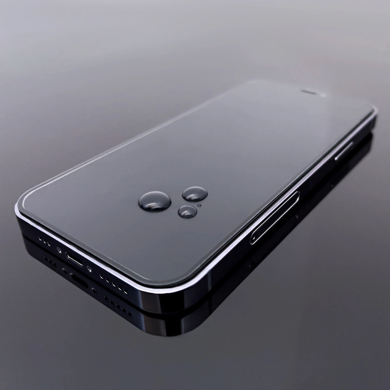 Na obrazie widoczny jest smartfon z przezroczystą folią ochronną na ekranie, na której znajdują się krople wody. Telefon leży na ciemnej, odbijającej powierzchni.