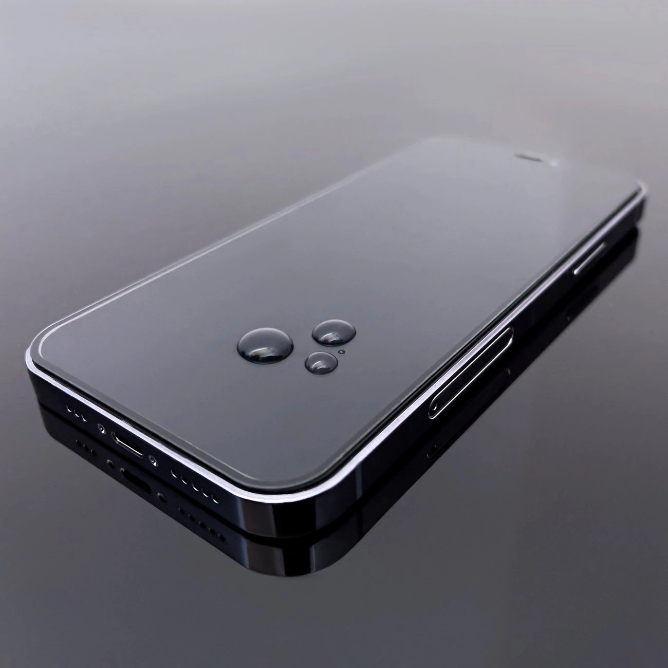 Na obrazie widoczny jest smartfon z nałożonym szkłem hartowanym, na którym znajdują się krople wody. Telefon leży na gładkiej, odbijającej powierzchni.
