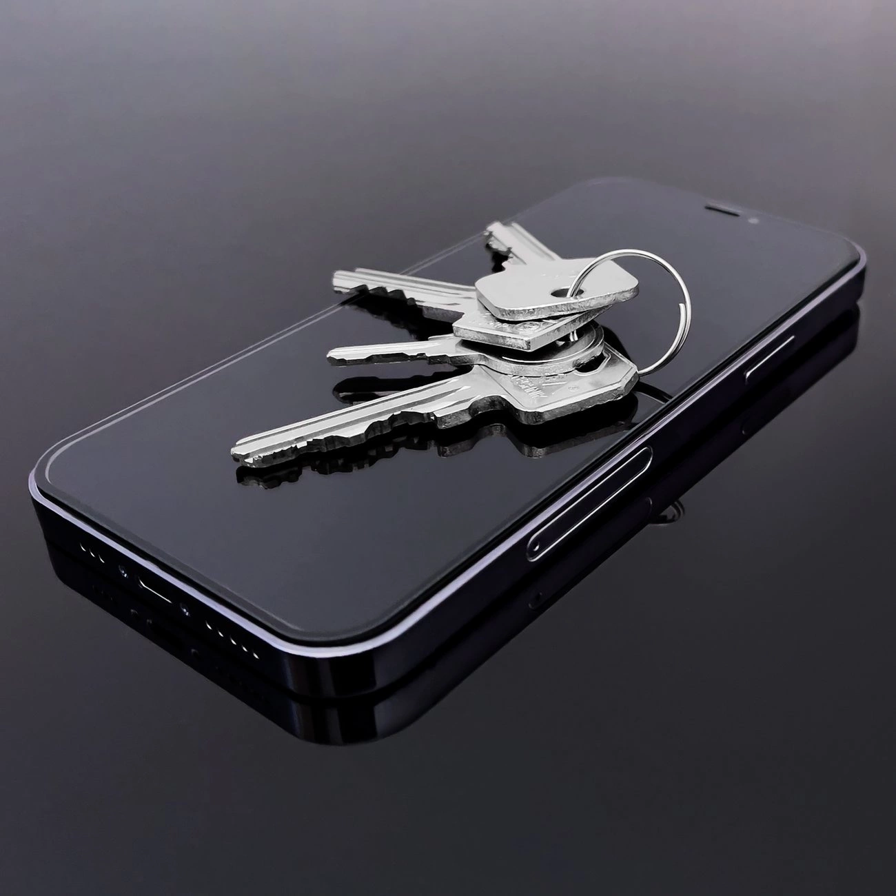 Na ciemnym tle leży smartfon z czarnym ekranem, na którym znajduje się kilka metalowych kluczy. Telefon ma widoczne przyciski boczne i porty na krawędzi.