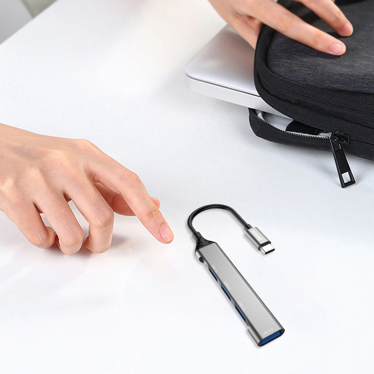 Na białym stole leży czarny hub USB-C z kilkoma portami USB-A. Obok znajduje się ręka oraz czarna torba z zamkiem błyskawicznym.