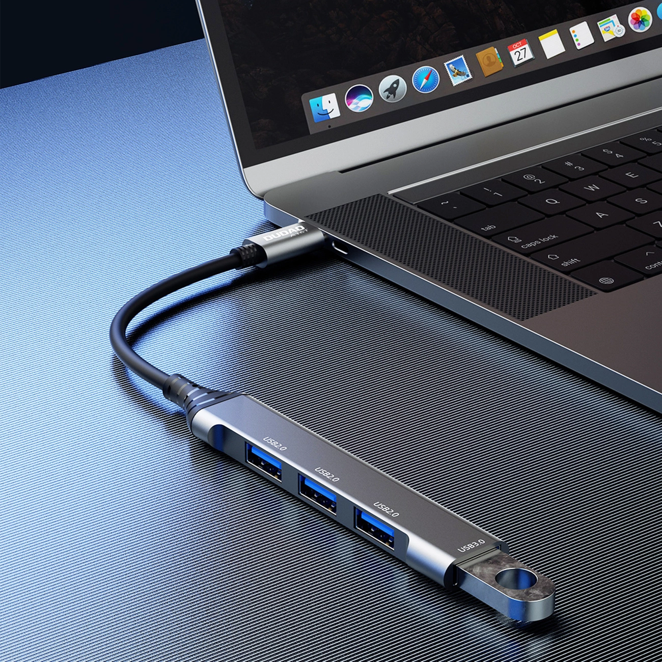 Na obrazie widać laptopa z podłączonym czarnym hubem USB-C, który posiada cztery porty USB-A oznaczone jako USB2.0 i USB3.0. Na kablu widoczne jest logo \'Dudao\'.
