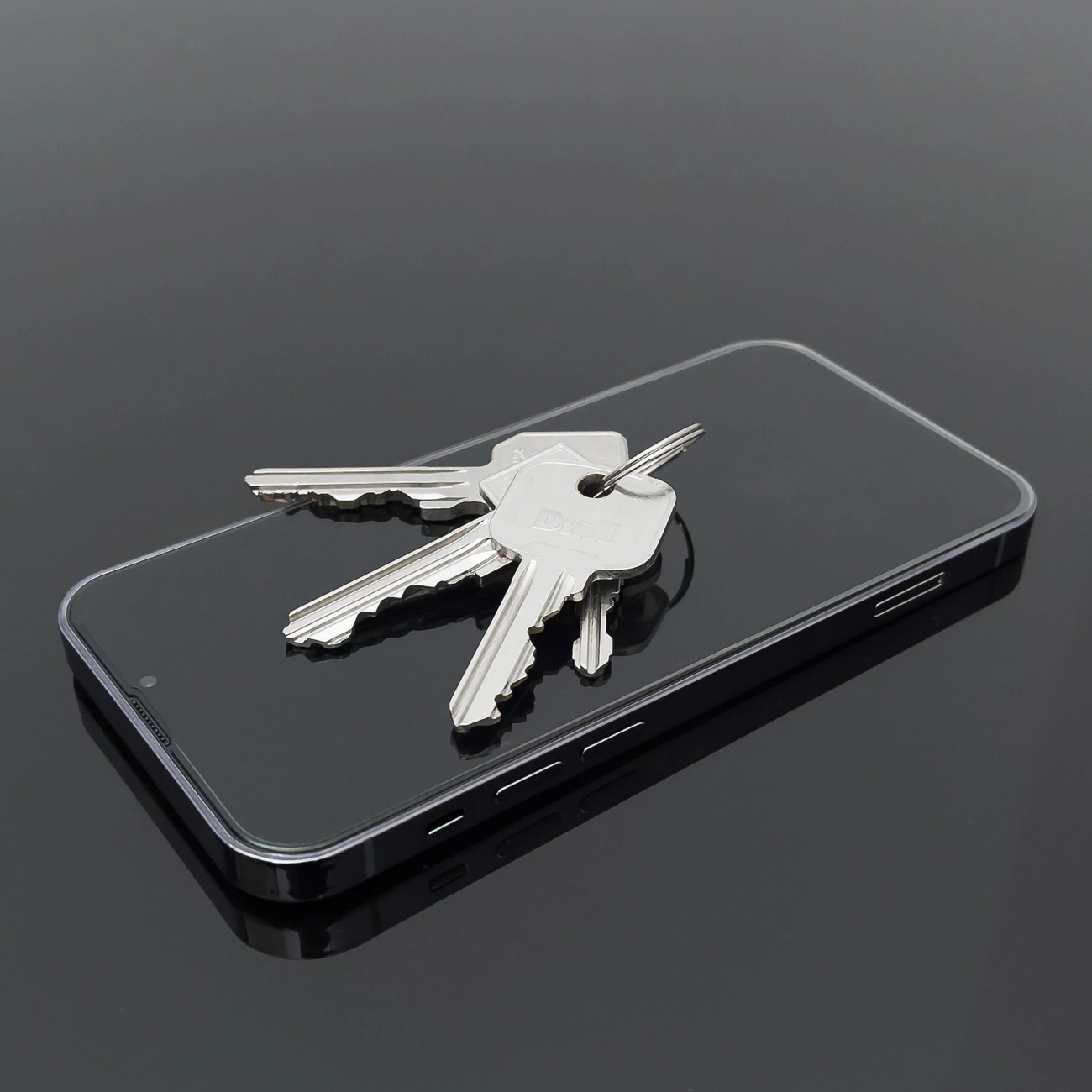 Na ciemnej powierzchni leży smartfon z kilkoma metalowymi kluczami na ekranie. Telefon ma ciemną obudowę i widoczne przyciski boczne.