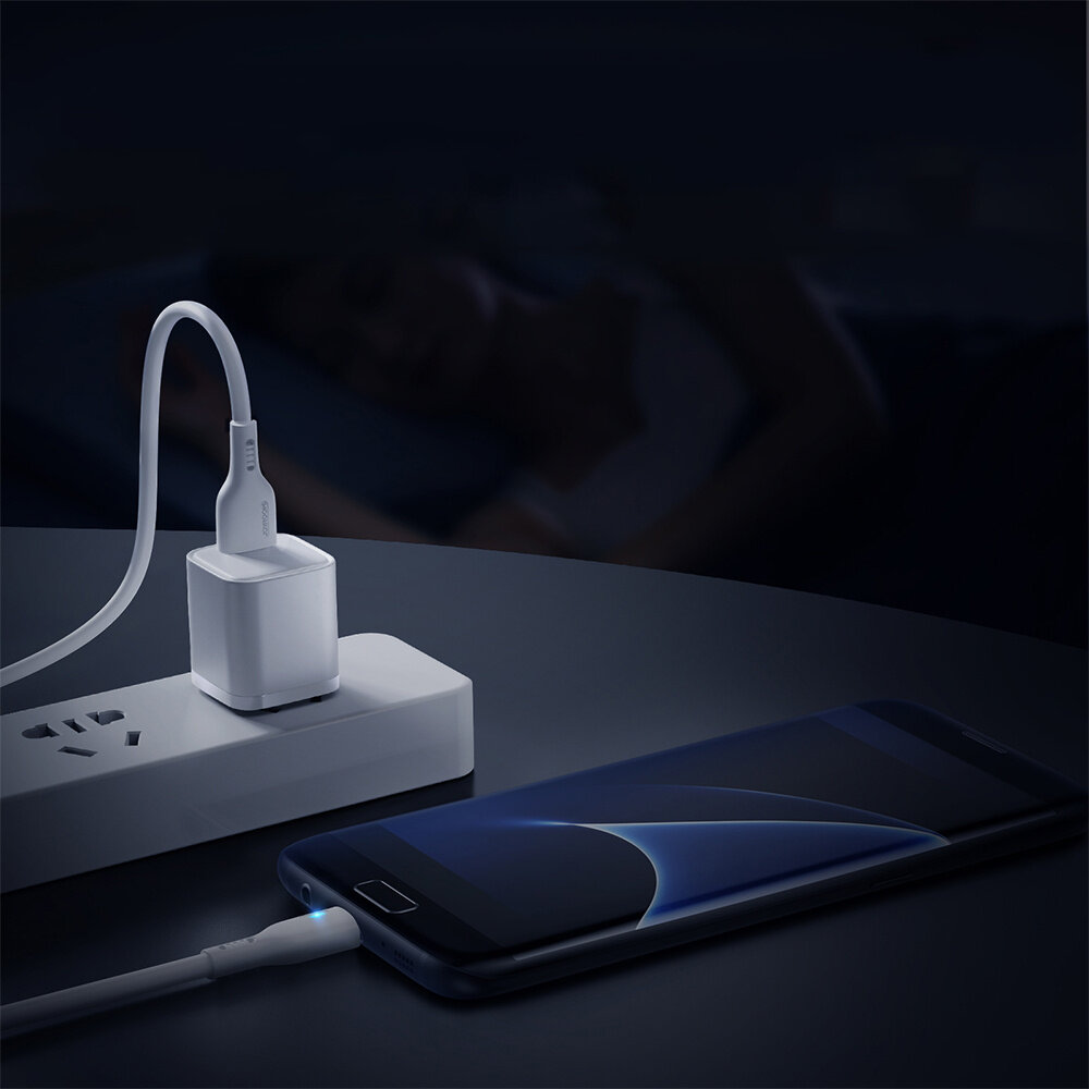 Na ciemnym tle widać telefon podłączony do ładowarki za pomocą białego kabla USB. W tle znajduje się osoba leżąca na łóżku.