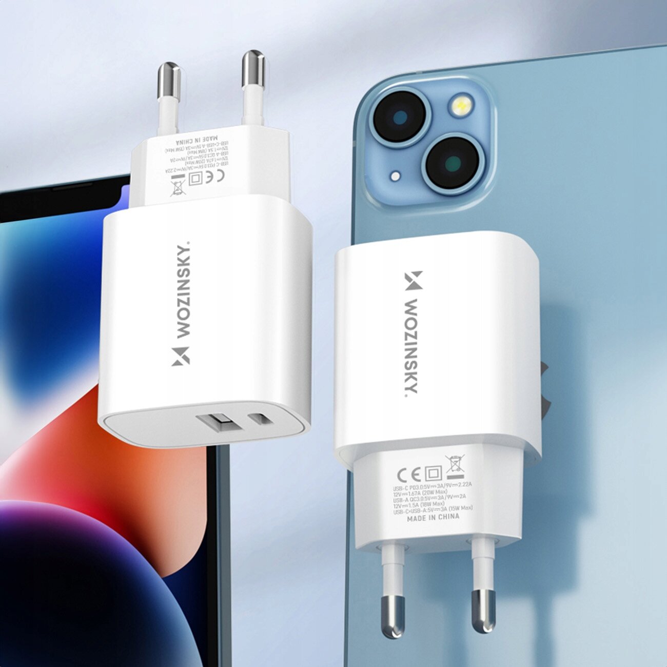 Na obrazie widoczne są dwie białe ładowarki sieciowe z logo \'WOZINSKY\', z wtyczkami typu USB-A i USB-C. W tle znajduje się fragment niebieskiego telefonu z dwoma aparatami.