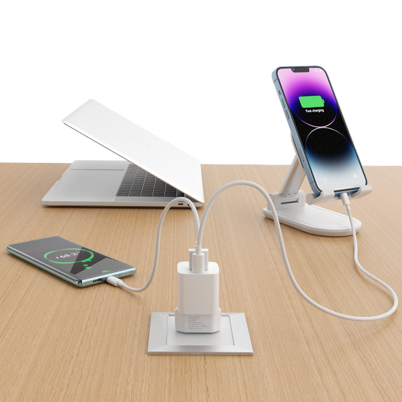 Na drewnianym stole znajduje się laptop, telefon na stojaku oraz drugi telefon leżący obok ładowarki sieciowej USB-C. Oba telefony są podłączone do ładowarki i wyświetlają ikony ładowania.
