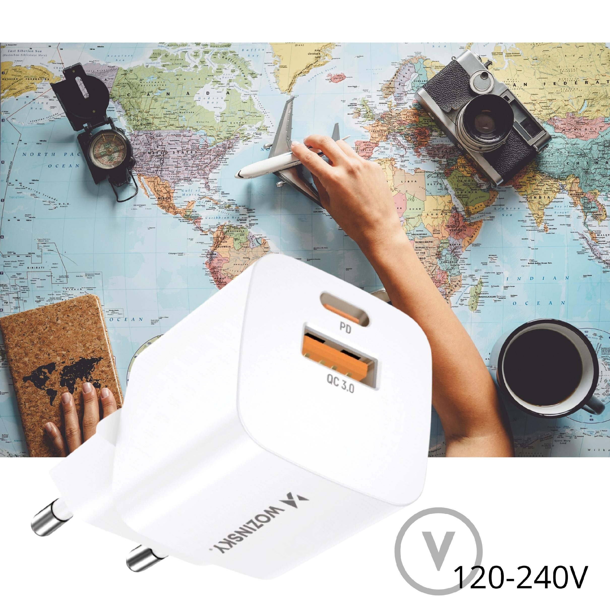 Na mapie świata leży biały adapter z logo \'Wozinsky\', posiadający porty oznaczone jako \'PD\' i \'QC 3.0\'. Obok widoczne są ręce trzymające model samolotu, aparat fotograficzny, kompas, notes z mapą oraz filiżanka z napojem.