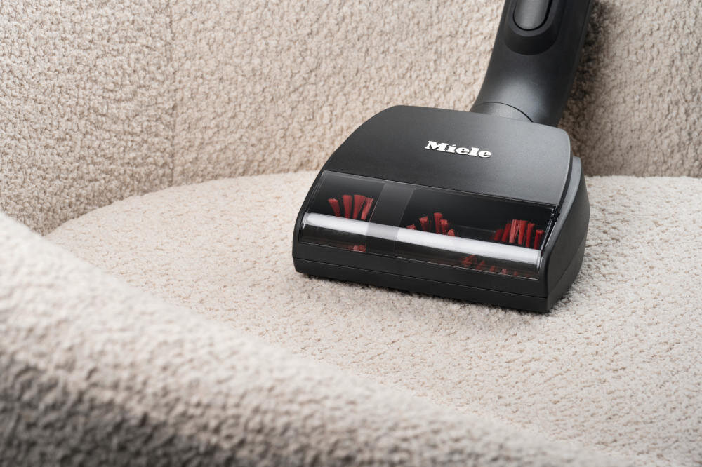 Czarna końcówka odkurzacza z widocznymi czerwonymi szczotkami na jasnym, teksturowanym materiale. Na końcówce widnieje logo \'Miele\'.