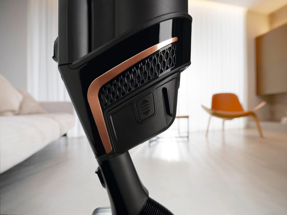 Zbliżenie na część odkurzacza z czarną obudową i miedzianym akcentem. W tle widoczny jest nowoczesny salon z jasną podłogą i meblami.