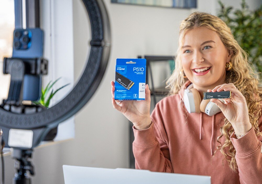 Dysk CRUCIAL P510 Kobieta z blond włosami uśmiecha się do kamery, trzymając opakowanie i sam dysk 'crucial P510', obok znajduje się statyw z telefonem, format, prędkość, odczyt, zapis, radiator