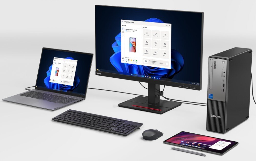 Na obrazie znajduje się zestaw komputerowy składający się z monitora, jednostki centralnej, klawiatury, myszy, laptopa i tabletu. Na ekranach widoczny jest interfejs systemu Windows 11, a na jednostce centralnej znajduje się logo Lenovo.