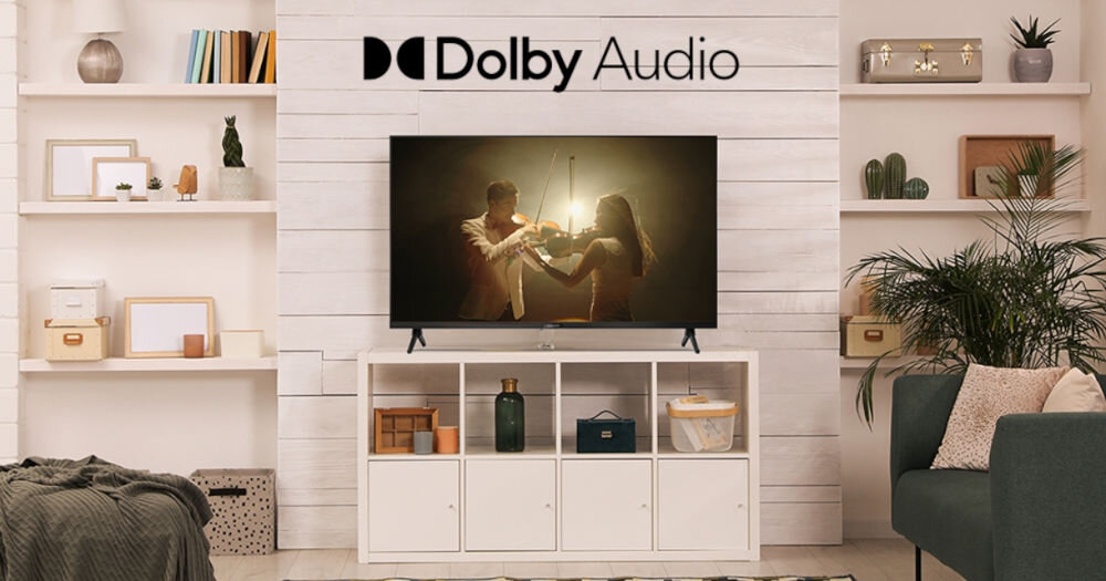 W salonie na białej szafce stoi telewizor, na którym wyświetlany jest obraz pary grającej na skrzypcach. Nad telewizorem widnieje napis \'Dolby Audio\'.