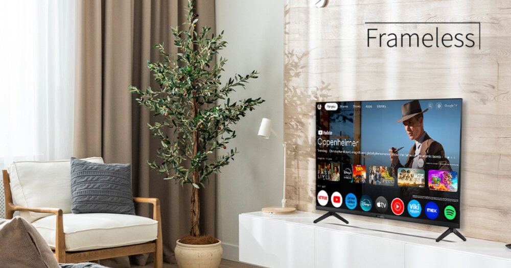 W salonie na białej szafce stoi telewizor z włączonym interfejsem Google TV, obok znajduje się roślina w doniczce i fotel. Na ścianie widoczny jest napis \'Frameless\'.