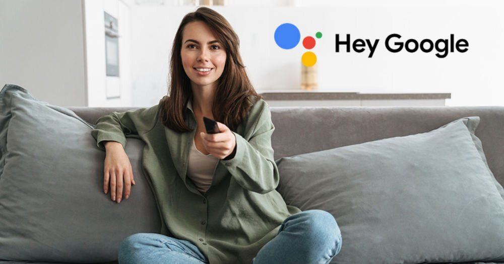 Osoba siedzi na kanapie trzymając pilot, obok widoczne jest logo Google z napisem \'Hey Google\'.