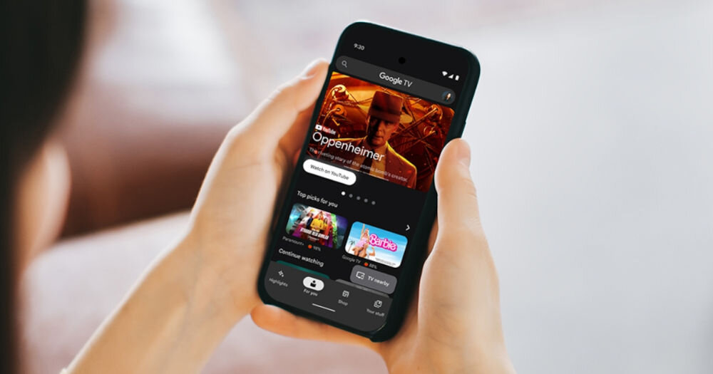 Osoba trzyma smartfon, na którym wyświetlana jest aplikacja Google TV z filmem \'Oppenheimer\' na ekranie. Widać także inne rekomendacje filmowe, takie jak \'Barbie\'.