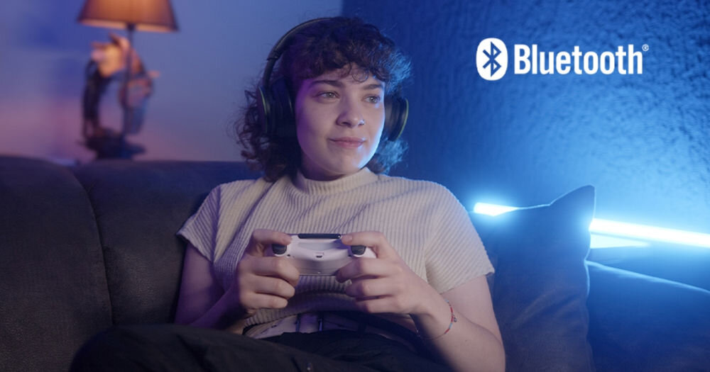 Osoba siedzi na kanapie z kontrolerem do gier, nosząc słuchawki. W tle widoczne jest logo \'Bluetooth\'.