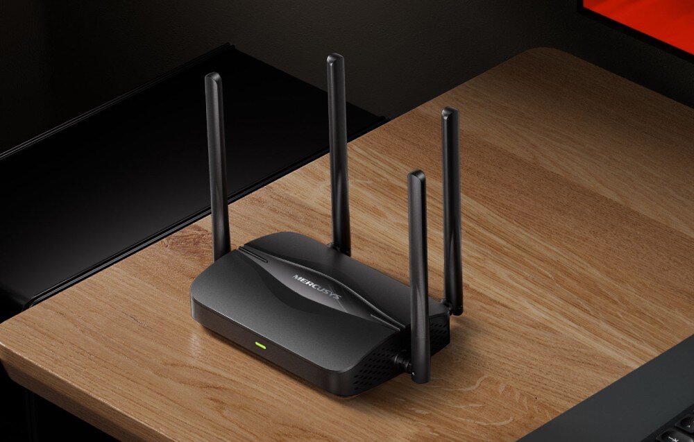 Router MERCUSYS MR25BE Czarny router bezprzewodowy z czterema pionowymi antenami ustawiony na jasnym drewnianym biurku w pobliżu klawiatury i monitora komputerowego