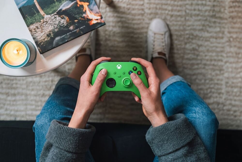 Osoba trzyma zielony kontroler do gier z widocznym logo Xbox. Obok znajduje się stolik z zapaloną świecą i książką z okładką przedstawiającą ognisko.