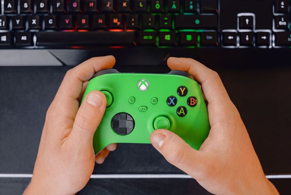 Osoba trzyma zielony kontroler do gier z widocznymi przyciskami X, Y, A, B oraz logo Xbox. W tle znajduje się klawiatura z podświetleniem RGB.