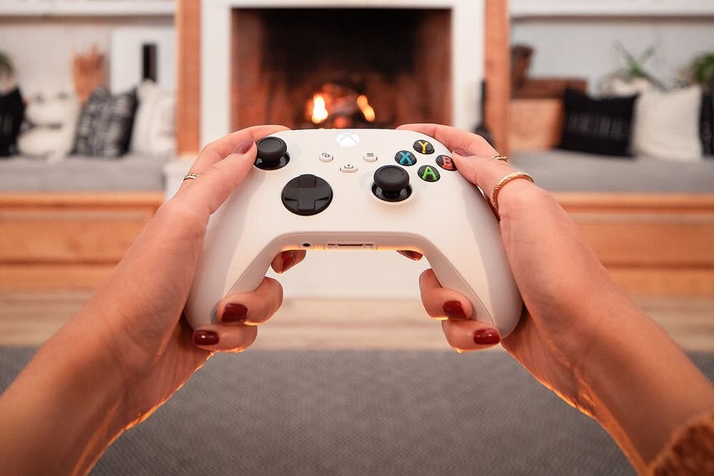 Osoba trzyma biały kontroler do gier z widocznymi przyciskami i joystickami. W tle znajduje się kominek z płonącym ogniem.
