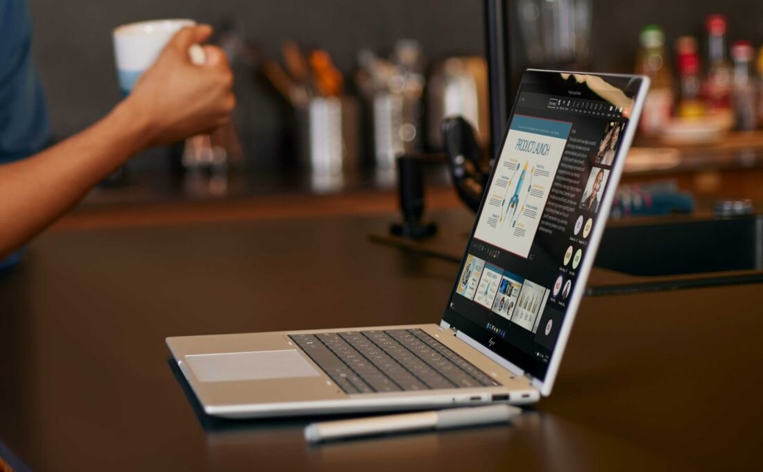 Laptop HP EliteBook X Flip w srebrnej obudowie na drewnianym stole w kawiarni, ekran z prezentacją biznesową zawierającą wykresy i dane analityczne, w tle osoba trzymająca kubek kawy, atmosfera pracy zdalnej.