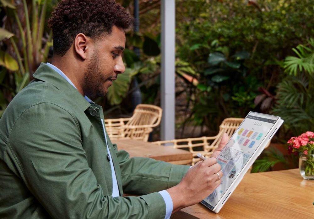 Mężczyzna w zielonej kurtce pracujący na laptopie HP EliteBook X Flip w trybie tabletu na drewnianym stole w ogrodzie, używa piórka cyfrowego do edycji kolorowych wykresów i diagramów, w tle kwiaty i rośliny doniczkowe. - Pamięć DDR5