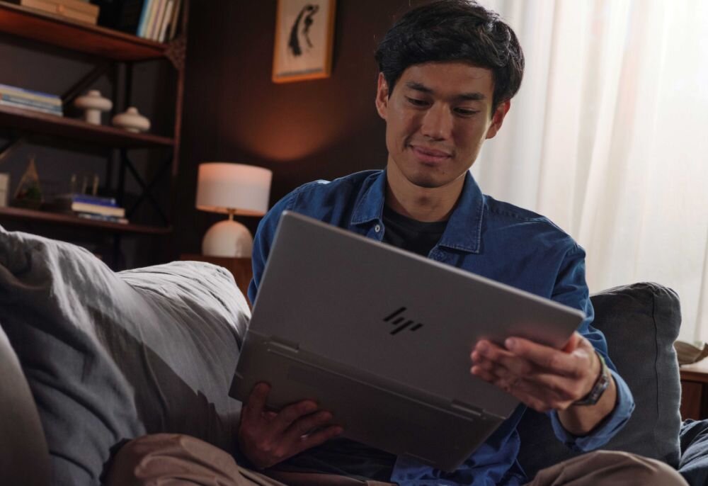 Młody mężczyzna w niebieskiej koszuli uśmiechający się podczas pracy na laptopie HP EliteBook X Flip w trybie tabletu, w przytulnym wnętrzu z półkami książek i ciepłym oświetleniem, podkreślający niski całkowity koszt posiadania. - Certyfikat TCO, Ekologia