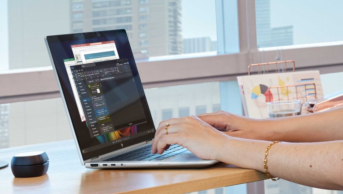 Osoba pracuje na srebrnym laptopie HP przy biurku w biurze z dużymi oknami; na ekranie otwarte są aplikacje biurowe, w tle widoczne wysokie budynki, na biurku leży myszka, szklanka z napojem oraz dokumenty z wykresami.