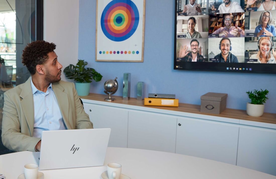 Mężczyzna w jasnej marynarce siedzi przy okrągłym stole z otwartym laptopem HP, patrzy na ścianę, na której wyświetlana jest wideokonferencja z wieloma uczestnikami; na stole filiżanki, w tle niebieska ściana z kolorowym obrazem, rośliny doniczkowe i segregatory. - polimerowy akumulator litowo-jonowy HP Long Life o pojemności 68 Wh