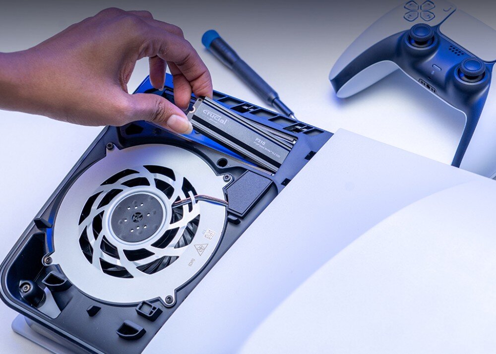 Dysk CRUCIAL P310 Ręka wkłada dysk SSD z napisem 'crucial P310' do gniazda w obudowie konsoli, obok leży kontroler i śrubokręt, pojemność, kompatybilność